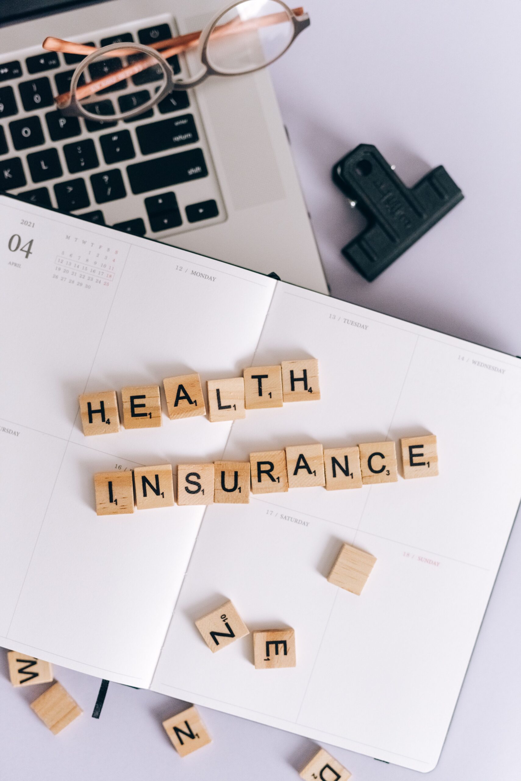 Scrabble tiles spelling 'Health Insurance' on a calendar, with laptop and stationery.